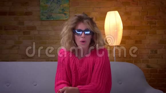 担心金发家庭主妇穿着粉红色毛衣和3D眼镜在舒适的家看恐怖电影视频的预览图