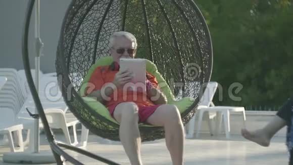 老人坐在挂椅上在酒店综合体放松可爱的小女孩站在爷爷身边休息视频的预览图