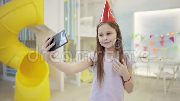 戴着生日帽的漂亮小女孩在镜头前的智能手机上自拍视频的预览图