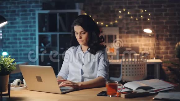 精疲力竭的年轻女士用笔记本电脑打字然后晚上在办公室打哈欠视频的预览图