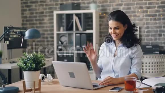 快乐的年轻女子一边说一边挥舞着手看着办公室里的笔记本电脑屏幕视频的预览图