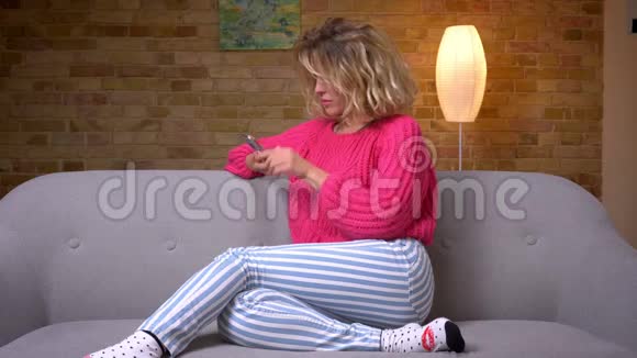 漂亮的家庭主妇穿着粉红色的毛衣坐在沙发上智能手机在家里的气氛中手摇着相机视频的预览图