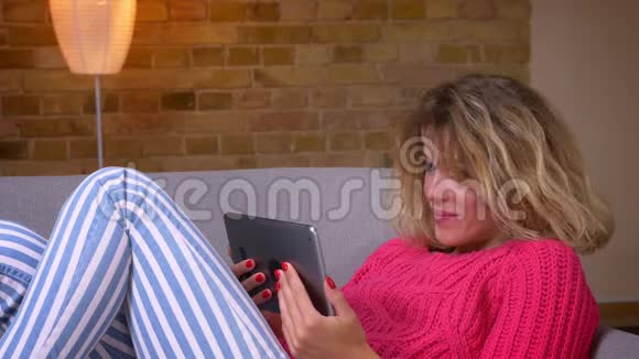 近景拍摄的金发家庭主妇穿着粉红色毛衣躺在沙发上在舒适的家庭氛围中在平板电脑上用视频聊天视频的预览图