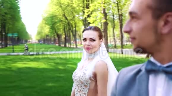 快乐的年轻新娘在时髦的新郎身边走来走去静静地站在阳光明媚的公园里视频的预览图