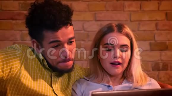 特写镜头非洲男人和高加索女孩坐在沙发上手提电脑在家欢乐地笑视频的预览图