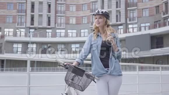 在城市建筑背景下戴着自行车头盔的可爱金发女人站在自行车上女士是视频的预览图