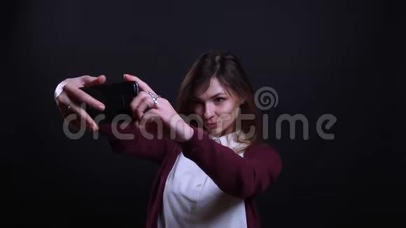 年轻的黑发女商人在黑色背景下愉快地在手机上制作漂亮的自拍照片的肖像视频的预览图