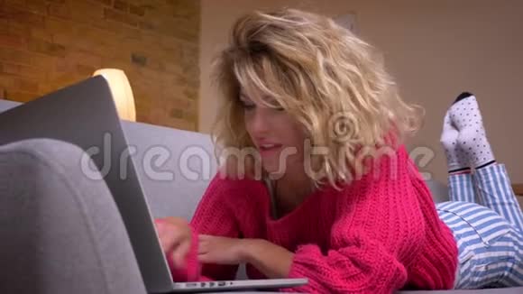 在舒适的家庭氛围中一位身穿粉色毛衣的金发家庭主妇躺在沙发上用笔记本电脑冲浪视频的预览图