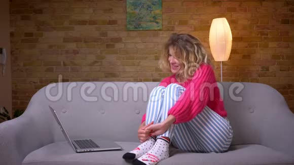 家庭主妇穿着粉红色毛衣的特写镜头拥抱她的膝盖在家里的气氛中手提电脑上有可视电话视频的预览图