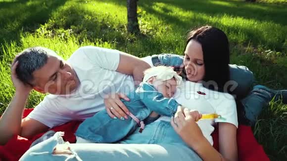 爱的父母和小女儿在阳光明媚的公园里的绿草上休息视频的预览图