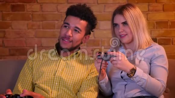 非洲男人和他的金发白种人女朋友在舒适的家中用操纵杆玩电子游戏视频的预览图
