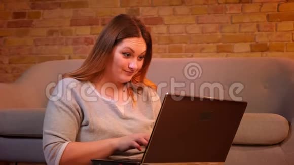漂亮的加码模特坐在地板上微笑着在笔记本电脑上打字在舒适的家庭氛围中为工作感到高兴视频的预览图