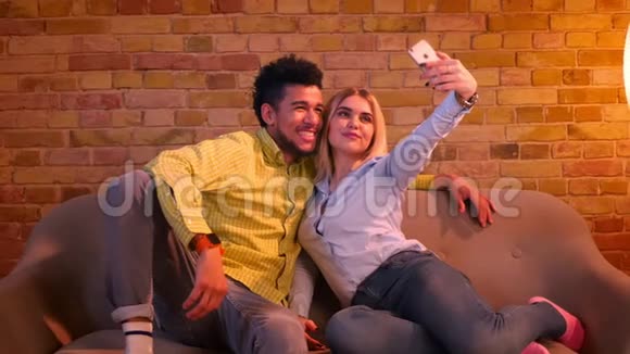 非洲男人和白种人女孩坐在沙发上在电话里自拍在家里一起快乐和美丽视频的预览图