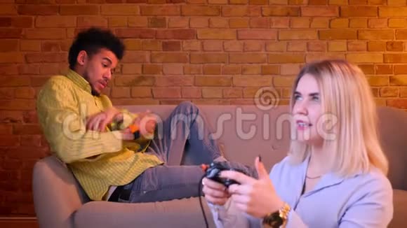 金发女孩聚精会神地在地板上玩电子游戏非洲男朋友在后台感到沮丧和孤独视频的预览图