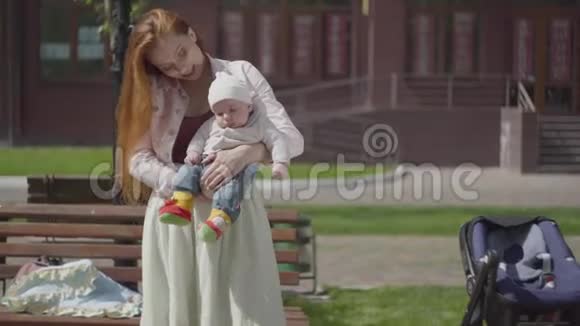 肖像漂亮的红发女人和坐在长凳上的孩子玩站在附近的婴儿车女士视频的预览图