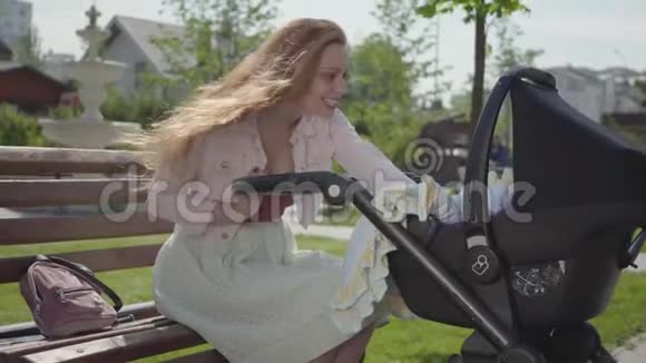 年轻快乐的红头发母亲坐在房子附近的长凳上坐着婴儿车微笑着在一个很好的地方和他说话视频的预览图