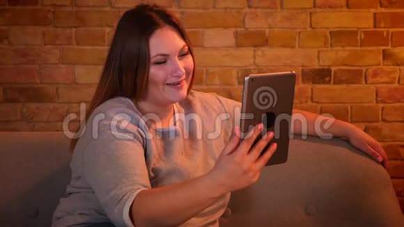 超重快乐的女性模特坐在沙发上在舒适的家庭氛围中平板电脑上有一个视频卡视频的预览图