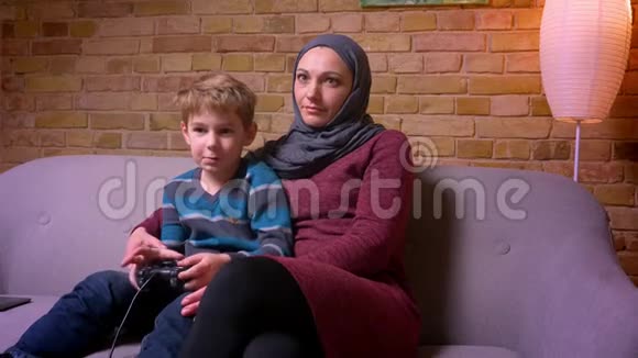 专注的小男孩在玩视频游戏他的穆斯林母亲戴着头巾试图拿起操纵杆试着玩视频的预览图