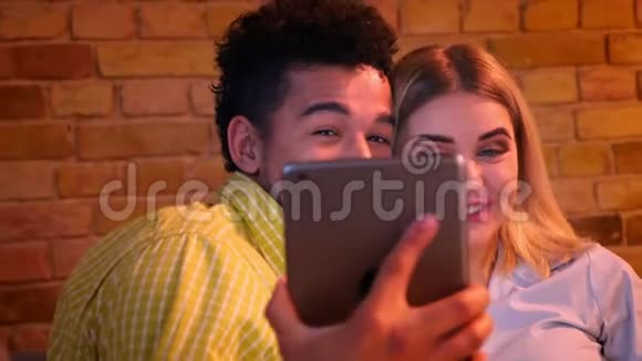 非洲男人和白种人女孩坐在沙发上在平板电脑上说着有趣的自拍照片在家里是快乐和快乐的视频的预览图