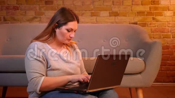 相当超重的学生坐在地板上微笑着在笔记本电脑上打字在舒适的家庭氛围中对工作感到满意视频的预览图