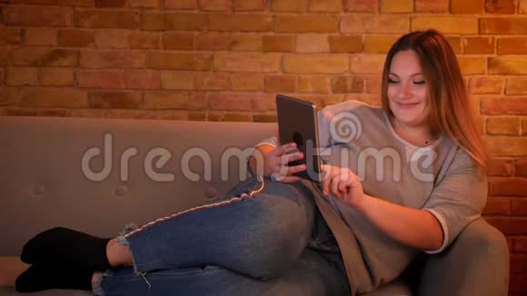 在舒适的家庭氛围中轻松加上大小的长发模特躺在沙发上专心地与平板电脑合作视频的预览图