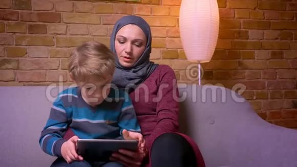集中的小男孩和他的穆斯林母亲坐在头巾上玩游戏在家里平板电脑的肖像视频的预览图