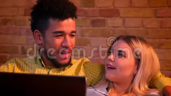特写镜头非洲男人和高加索女孩坐在沙发上手提电脑在家愉快地交谈视频的预览图