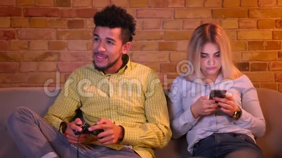 非洲男人用操纵杆玩电子游戏他的金发白种人女朋友在舒适的家里与智能手机合作视频的预览图
