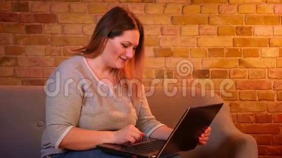在舒适的家庭氛围中长头发的超重学生坐在沙发上专心地在笔记本电脑上打字视频的预览图