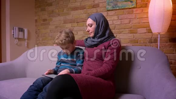 受虐的小男孩在平板电脑上玩游戏他的穆斯林母亲戴着头巾在家里观察他的活动视频的预览图