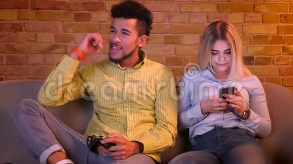 非洲男人专心地用操纵杆玩电子游戏他漂亮的白种人女朋友在舒适的环境中看电话视频的预览图