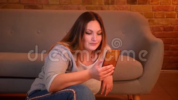 放松的加码长发模特在舒适的家居氛围中愉快地看着智能手机坐在地板上视频的预览图