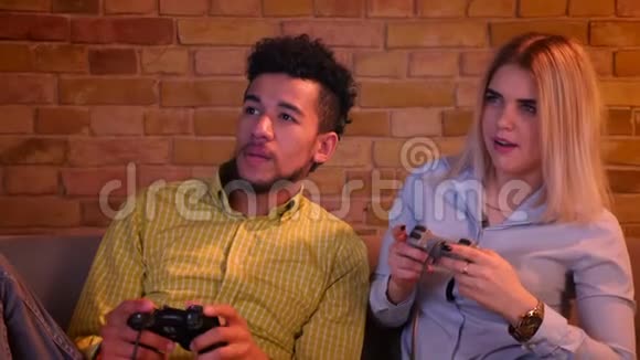 年轻的非洲男人和他的金发白种人女朋友在舒适的家庭氛围中玩游戏与操纵杆视频的预览图