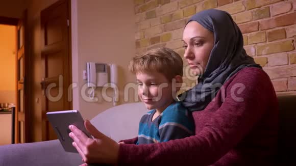 小男孩和他的穆斯林母亲戴着头巾在平板电脑上看电影的肖像在家里感到兴奋和有趣视频的预览图