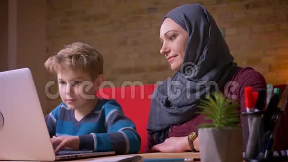 小男孩在笔记本电脑上玩游戏他的穆斯林母亲戴着头巾坐在附近观察他的活动视频的预览图