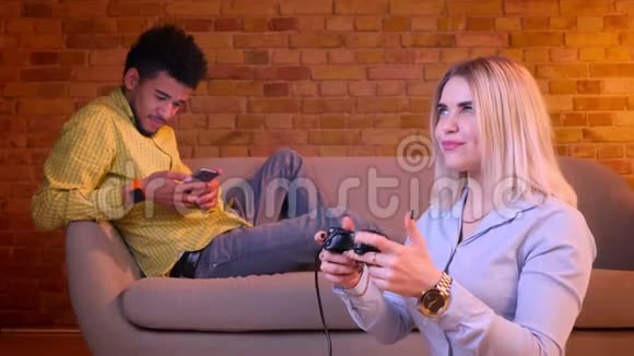 金发女孩在地板上玩视频游戏她的非洲男友在后台与智能手机合作视频的预览图