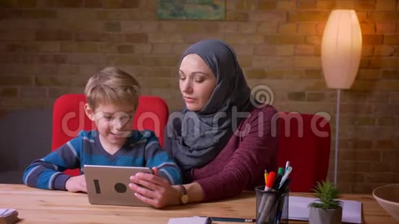 小男孩和他的穆斯林母亲戴着头巾一起在平板电脑上看动画片并讨论它们的肖像视频的预览图