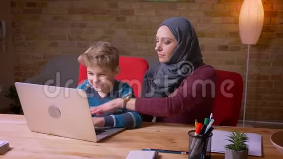 小男孩在笔记本电脑上打字他的穆斯林母亲戴着头巾坐在附近观察他的活动视频的预览图