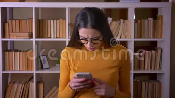 戴着智能手机眼镜的漂亮黑发老师的肖像转向相机在图书馆微笑视频的预览图
