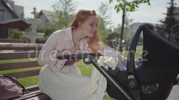 可爱的红发女人微笑着看着公园里坐在长凳上的婴儿车这位女士享受着阳光明媚的一天视频的预览图