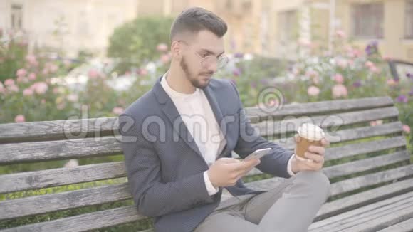 自信的年轻商人坐在户外长凳上的肖像看着他的智能手机和打字信息视频的预览图