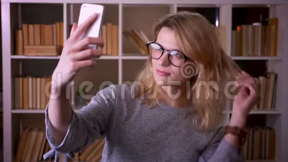 戴眼镜的漂亮中年金发老师在图书馆用智能手机做自拍视频的预览图