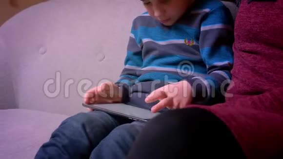 专心致志的小男孩在平板电脑上玩游戏他的穆斯林母亲戴着头巾坐在家附近视频的预览图