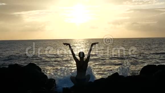 日落时分一位女士坐在岩石上观察海浪并在电影中举起手臂对抗海浪的剪影视频的预览图