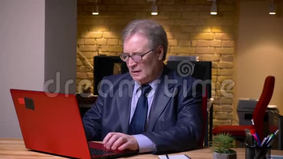 戴眼镜穿着正式服装在笔记本电脑上打字的老人在办公室里陷入尴尬的肖像视频的预览图
