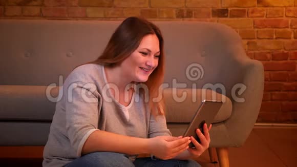 超重的长发女模特坐在地板上在平板电脑上打着电视聊天在舒适的家庭氛围中欢笑视频的预览图