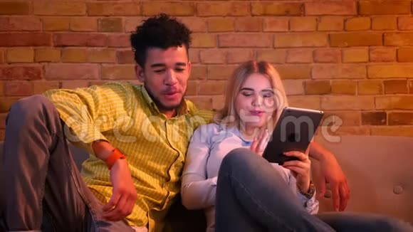 非洲男人和白种人女孩坐在沙发上拿着平板电脑在家里开心地笑着视频的预览图