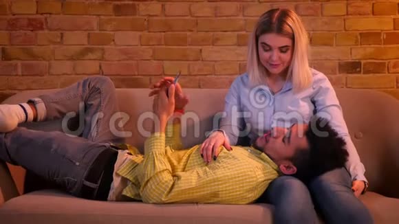 在家里的气氛中一个用智能手机工作的非洲男人躺在他高加索女朋友的腿上视频的预览图