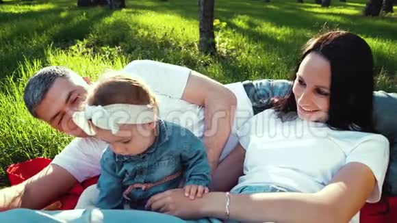 快乐的女婴和父母一起享受阳光明媚的天气躺在公园的毯子上视频的预览图