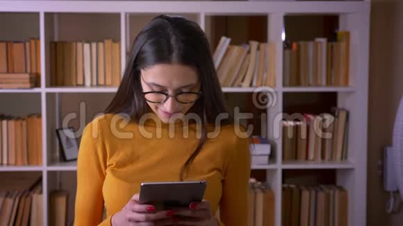 一位戴着眼镜手持平板电脑的漂亮黑发教师的画像在图书馆里对着镜头微笑视频的预览图